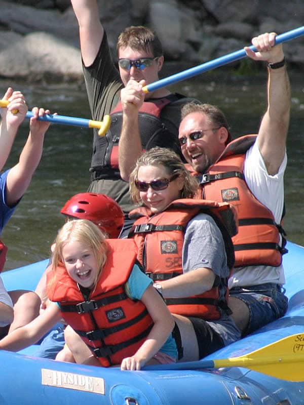 A family including a man a woman and a young girl are laughing as they are rafting down the Gunnison River. They are wearing life jackets and holding paddles as they raft down white water rapids.