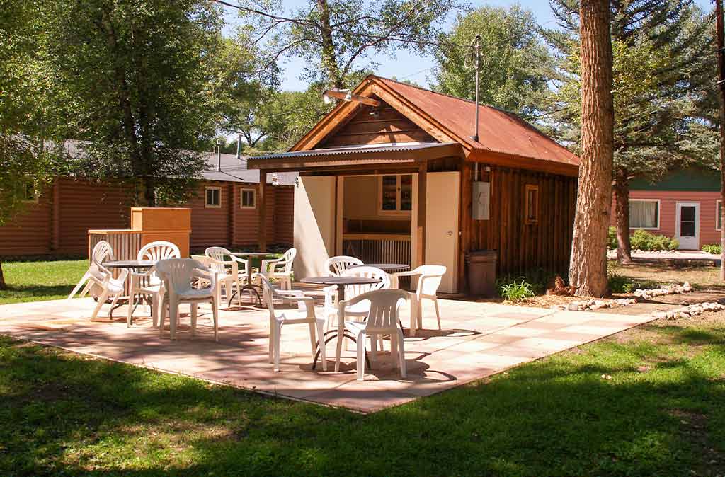 A patio seating area. There is a small shack with inside space and a large brick patio with chairs and tables, surrounded by beautiful grass and trees.