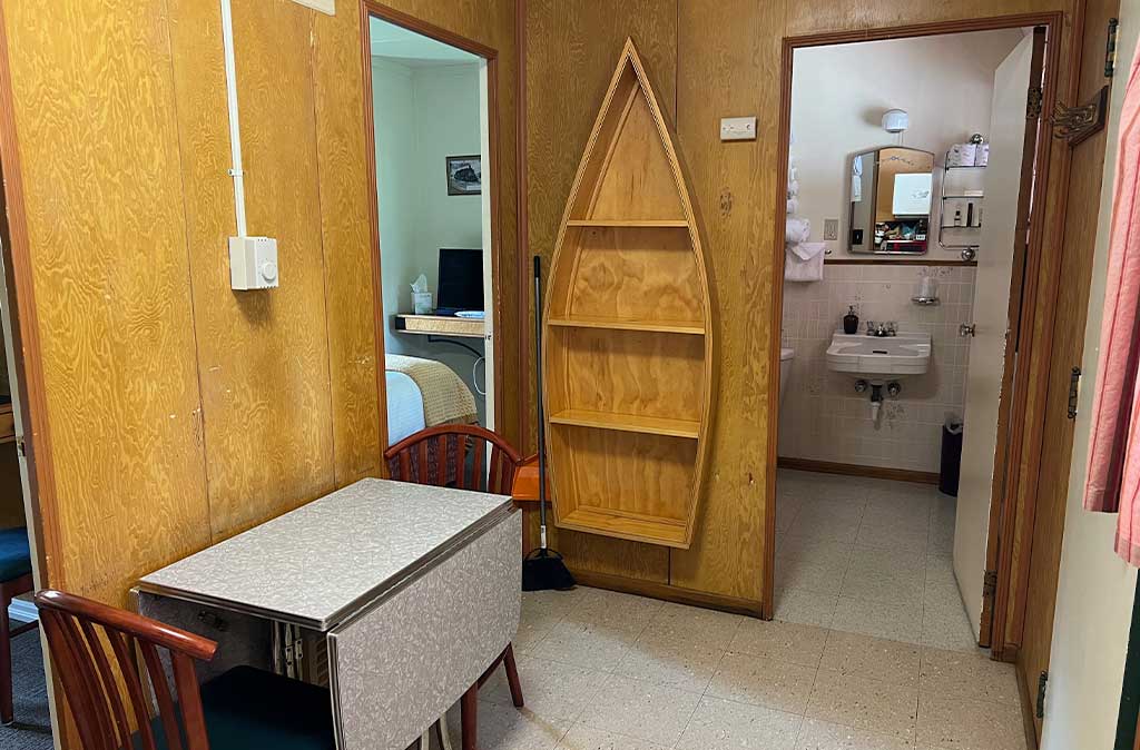 Photo of two-bedroom kitchenette unit at Island Acres Resort Motel showing vintage boat bookshelf, seating for two, and bathroom sink through the bathroom door.