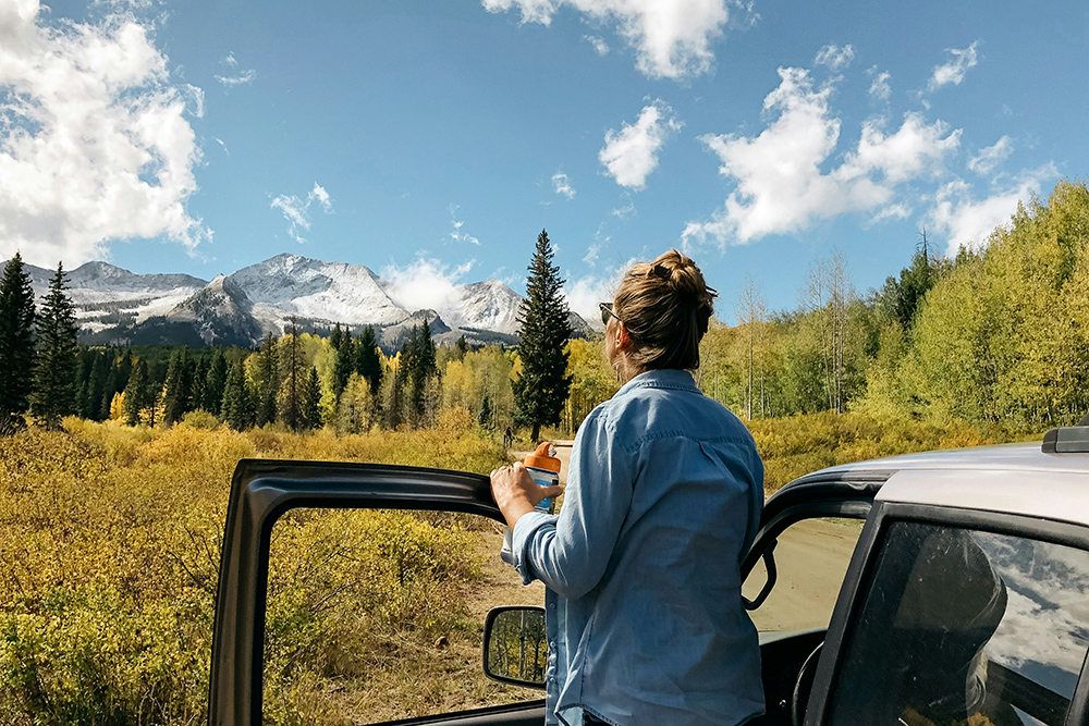 Photo of woman standing up outside driver's side car door looking out at a view of the Colorado Rocky Mountains covered in golden aspen and evergreen pine trees for Island Acres Resort blog on the 3 Best Drives for Colorado Leaf Peeping Near Gunnison